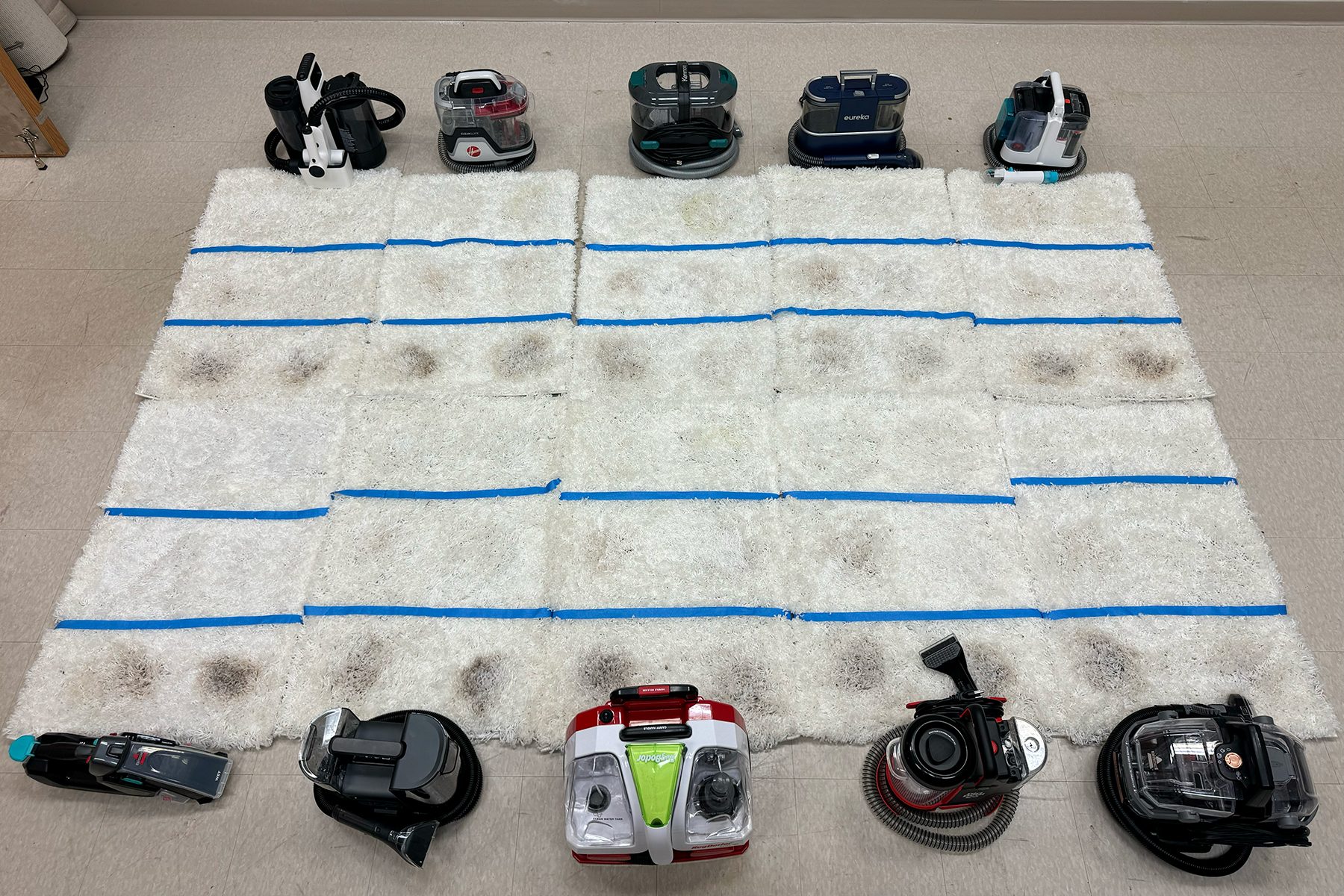 Carpet cleaner devices surround a dirt-stained white mat, arranged in rows on a tiled floor, indicating a cleaning comparison setup.