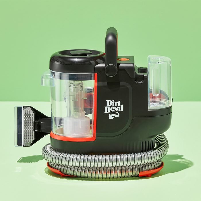 A compact vacuum cleaner resting on a light green surface, with a coiled hose at its base, labeled 