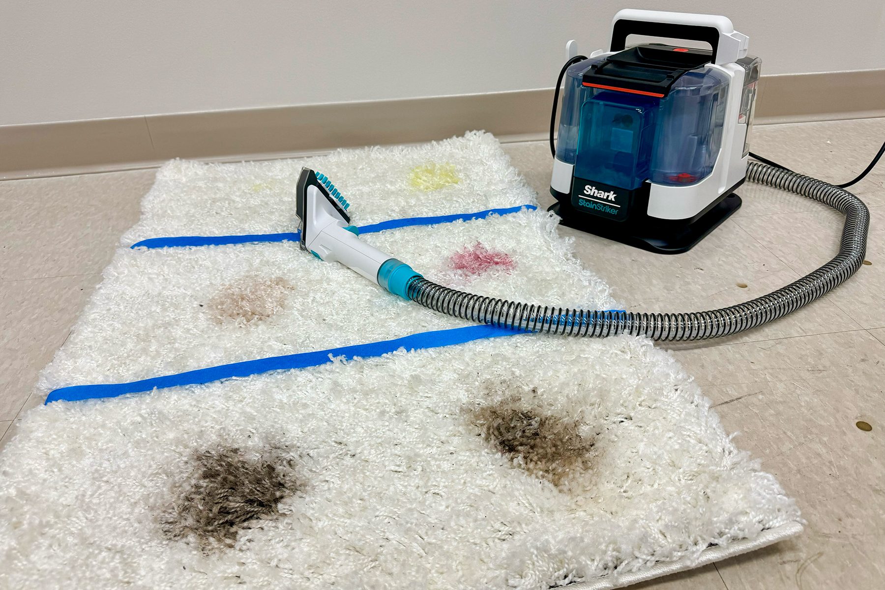 Carpet cleaning device resting on a stained carpet, surrounded by blue tape, in a room with a plain wall and floor tiles.
