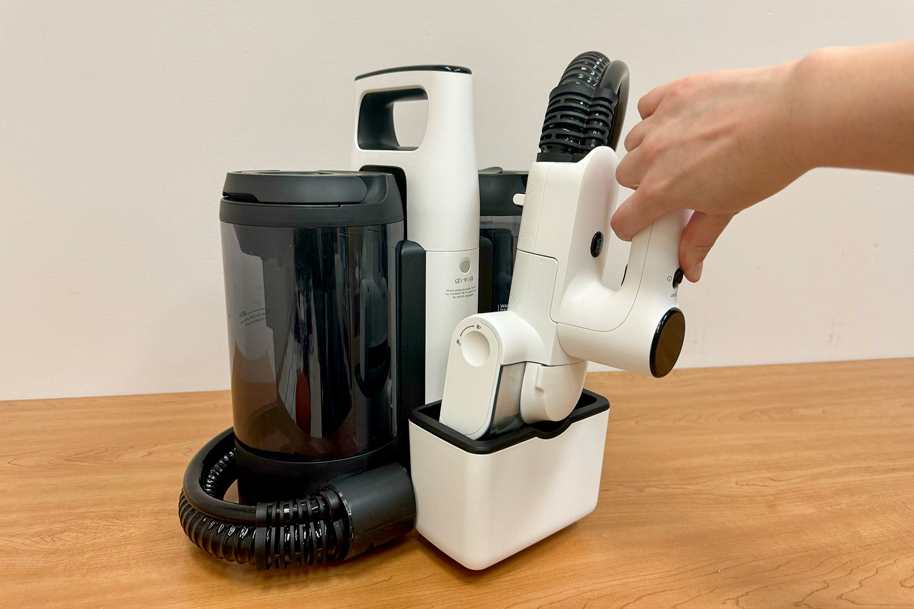 A handheld massaging device is being placed in its charging cradle on a wooden table, next to a black and white charging station.