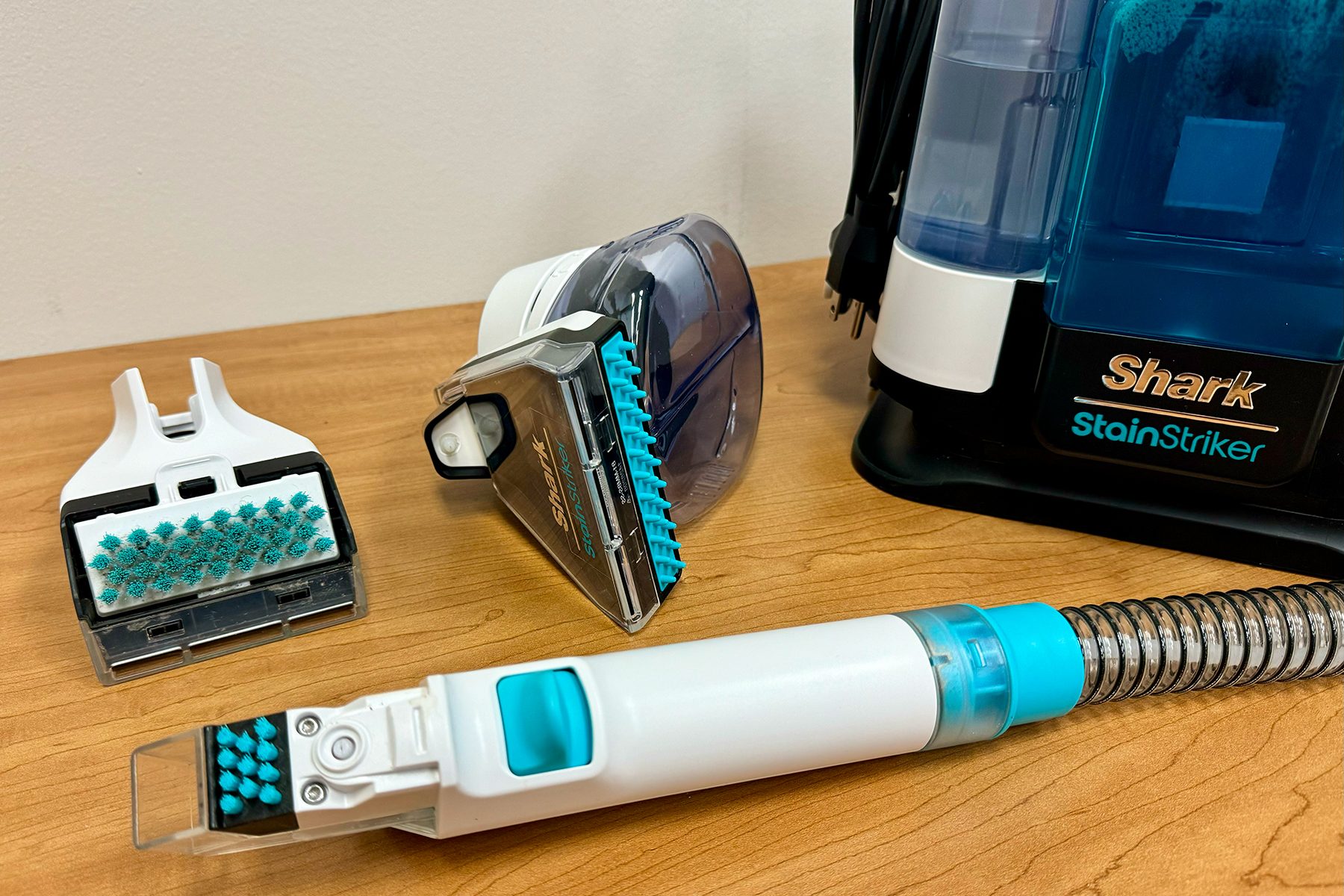 Vacuum attachments sit on a wooden table beside a Shark StainStriker machine, displaying various cleaning heads and a coiled hose.