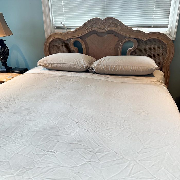 Bed with beige sheets and two pillows sits under a window with closed blinds; a lamp and table are nearby.