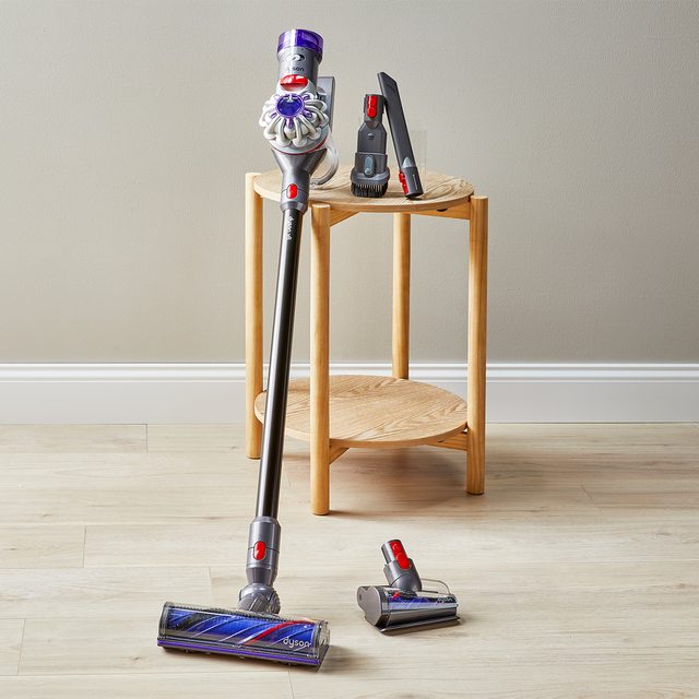 Vacuum cleaner stands on wooden floor with accessories placed on a small wooden table nearby, against a plain wall.