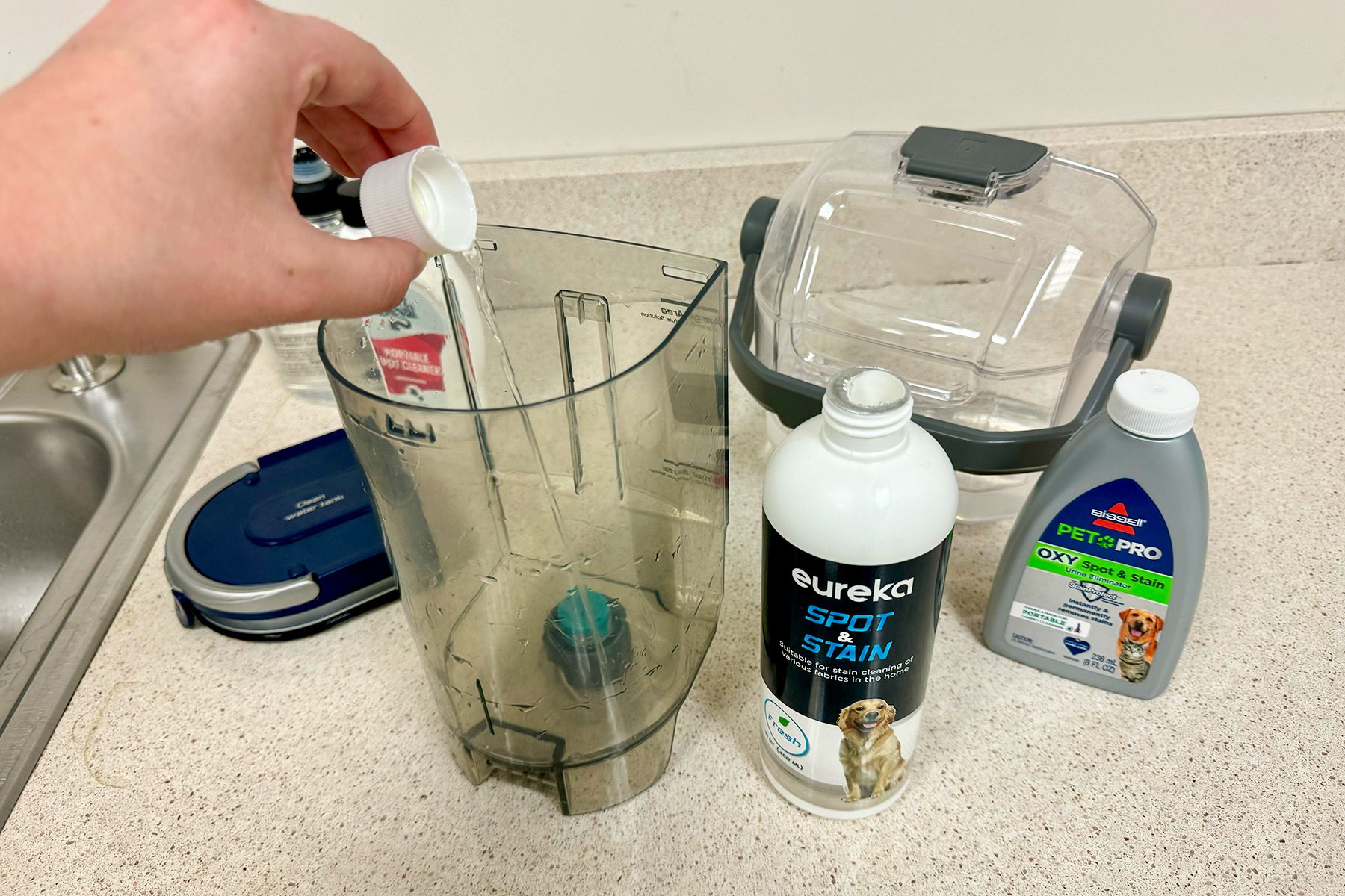 Hand pours cleaning solution into clear canister on countertop; nearby are bottles labeled "Eureka Spot & Stain" and "Bissell Pet Pro Oxy."