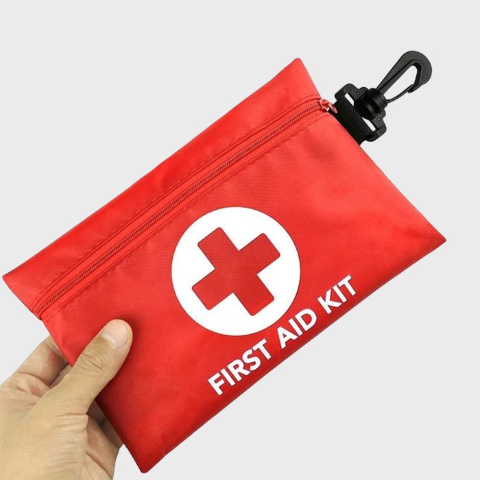 Red first aid kit held in hand, features a white cross and text, with a black clip attached, against a plain background.