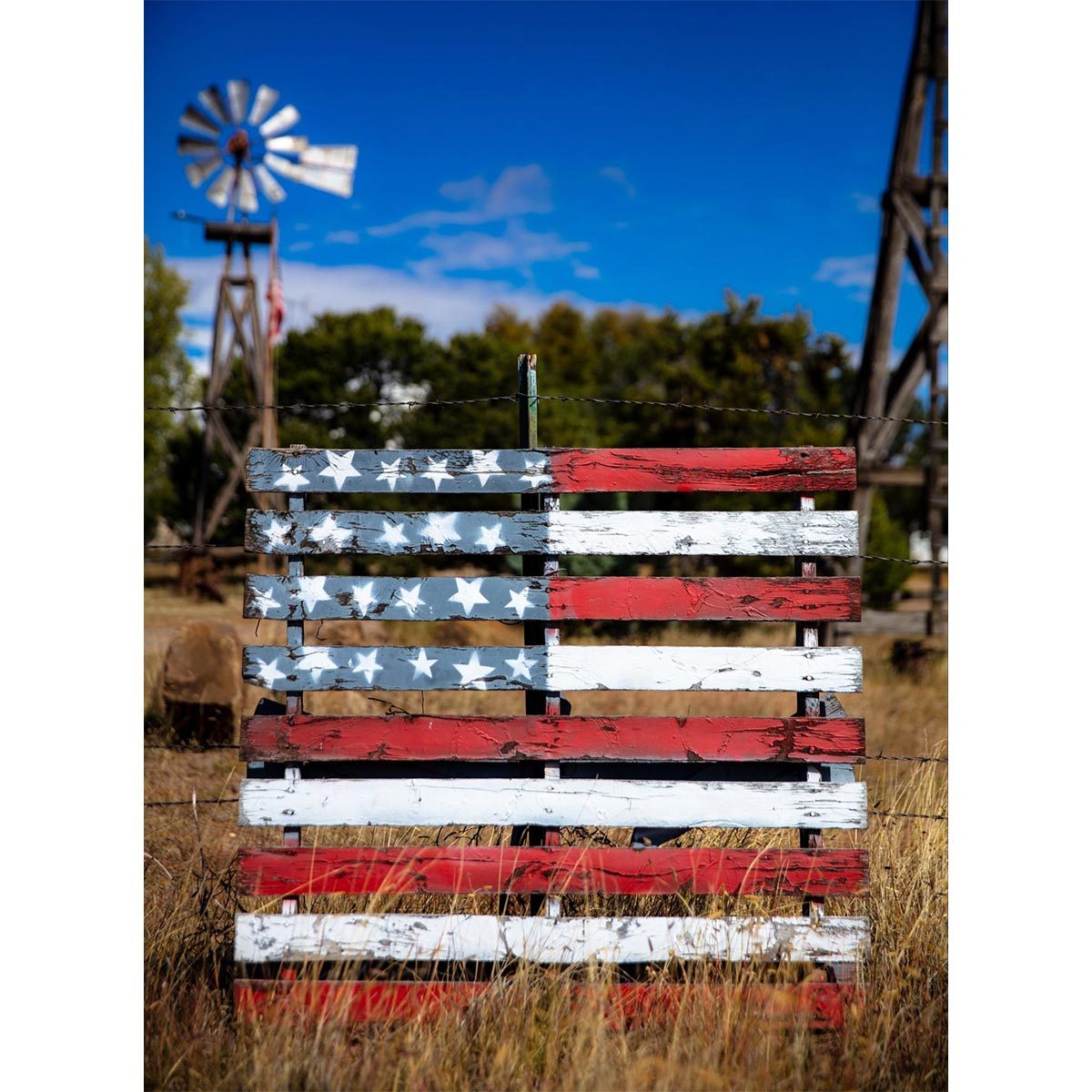 Painted wooden pallet resembling an American flag, leaning against a wire fence, with a windmill and trees in the grassy background.