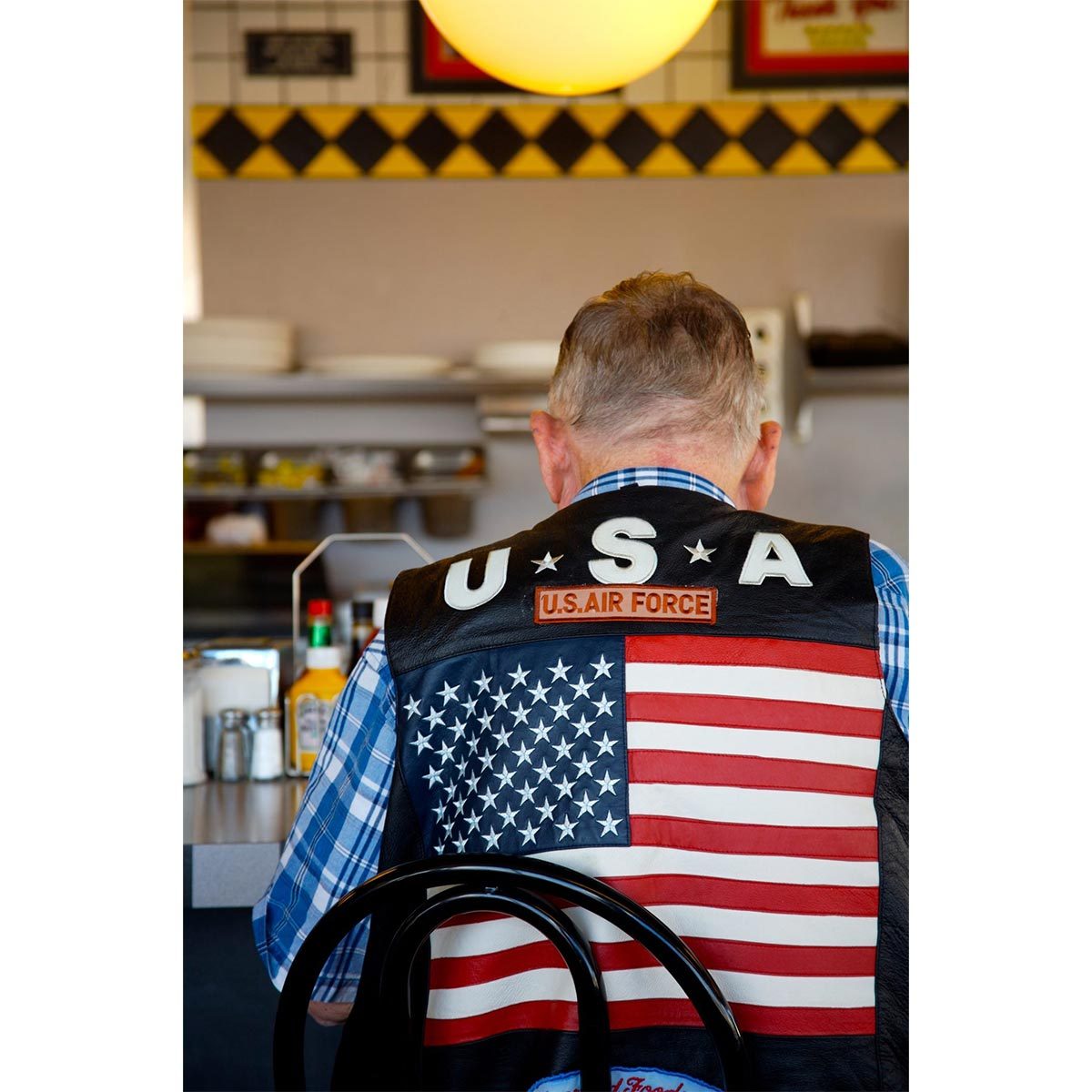 Man sits at diner counter, wearing vest displaying "USA," "U.S. Air Force," and a large American flag, with condiments nearby.