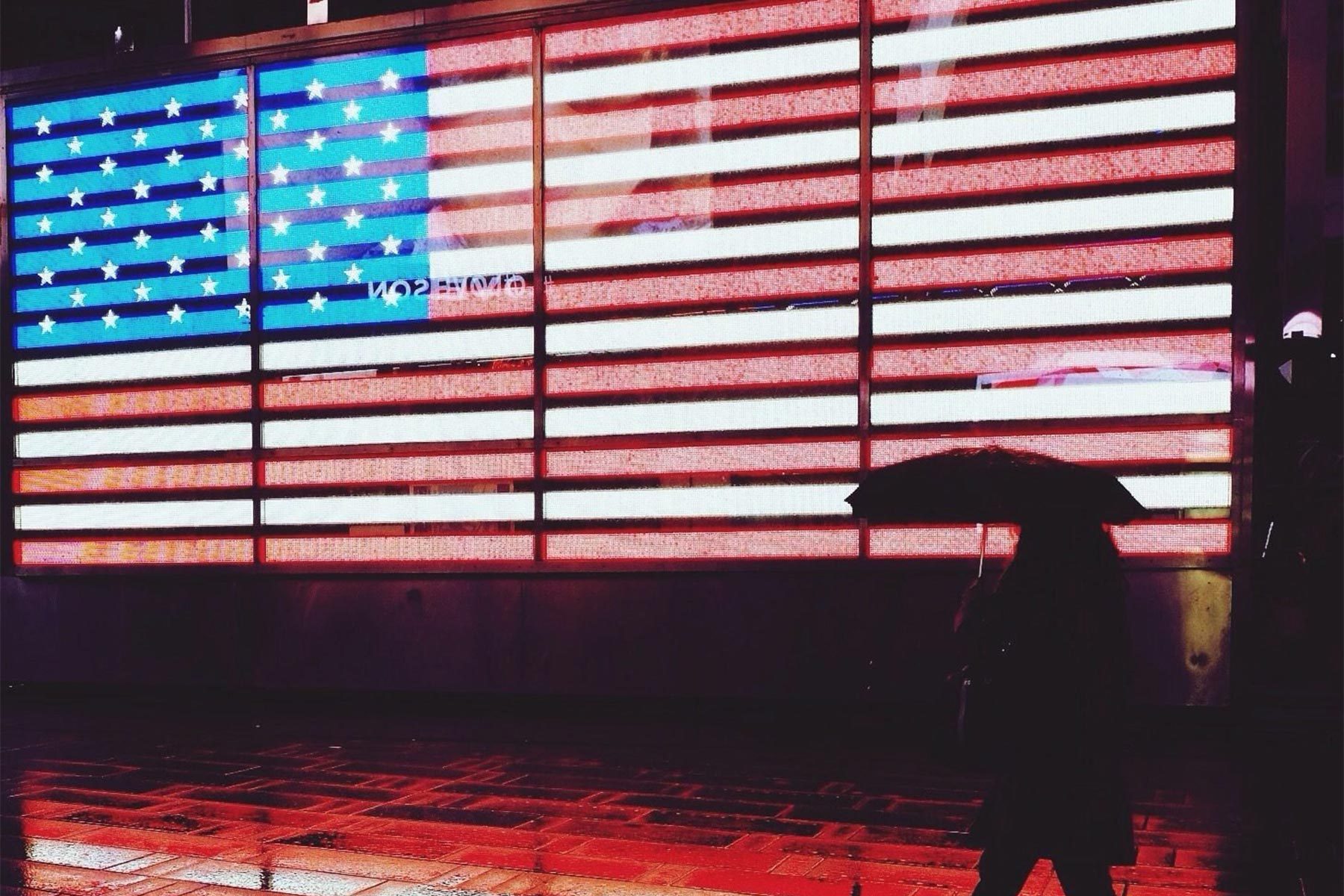Silhouette holding umbrella walks past large illuminated American flag display at night, with reflective wet pavement below.