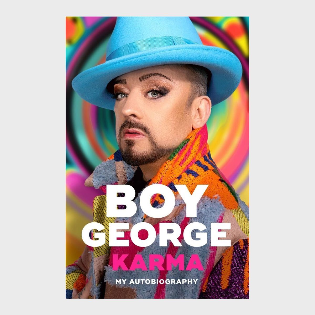 A person wears a blue hat and colorful coat, posing against a vibrant background. Text reads: "BOY GEORGE KARMA MY AUTOBIOGRAPHY."