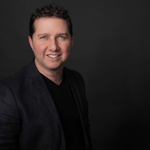 A person smiling slightly, wearing a dark blazer and shirt, set against a plain dark background.