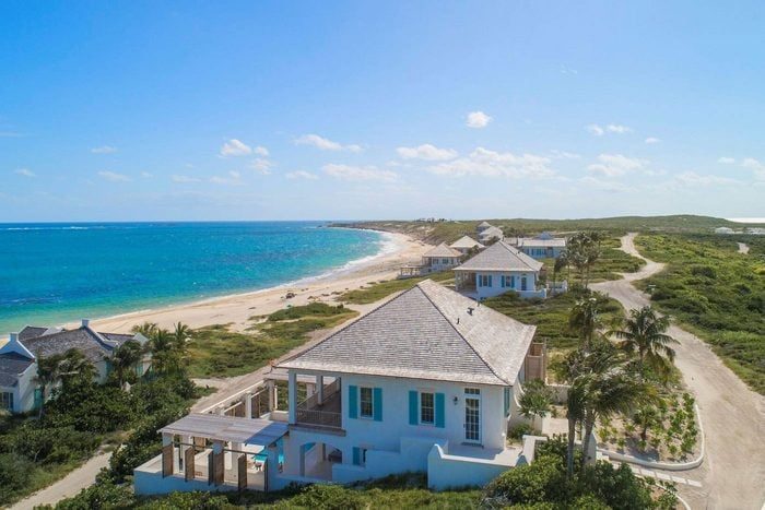 House overlooks ocean, surrounded by lush vegetation and other homes, with a sandy beach stretching alongside the turquoise water under a clear blue sky.