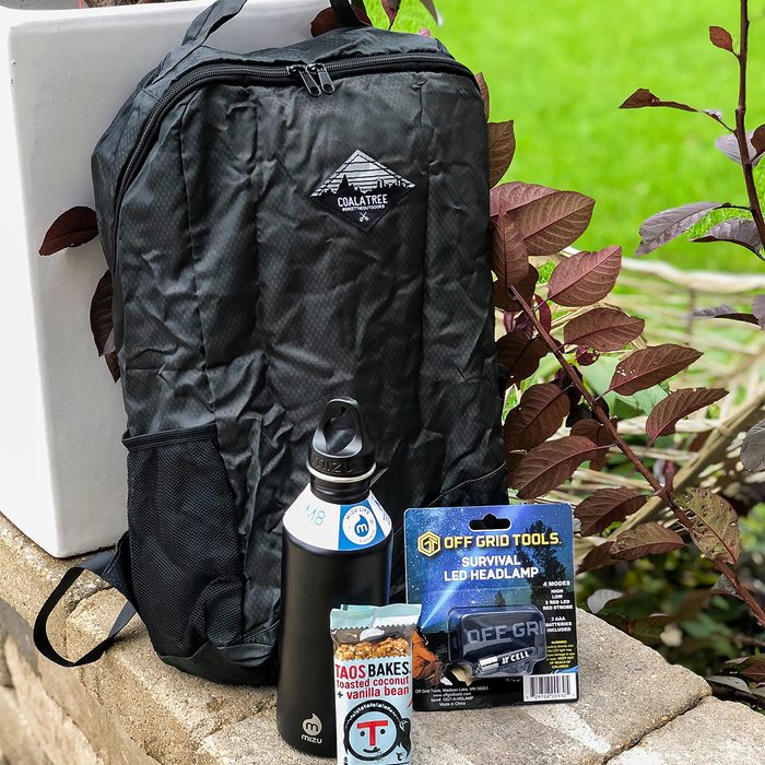 Backpack rests near foliage; bottle, snack, and headlamp package stand nearby, creating an outdoor-ready set. Text includes 