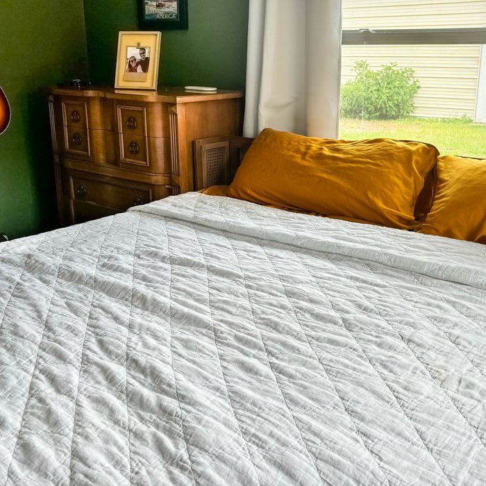 Bed with a white quilt, positioned beside a wooden dresser, yellow pillows, framed photos, and a green-walled, window-lit room.