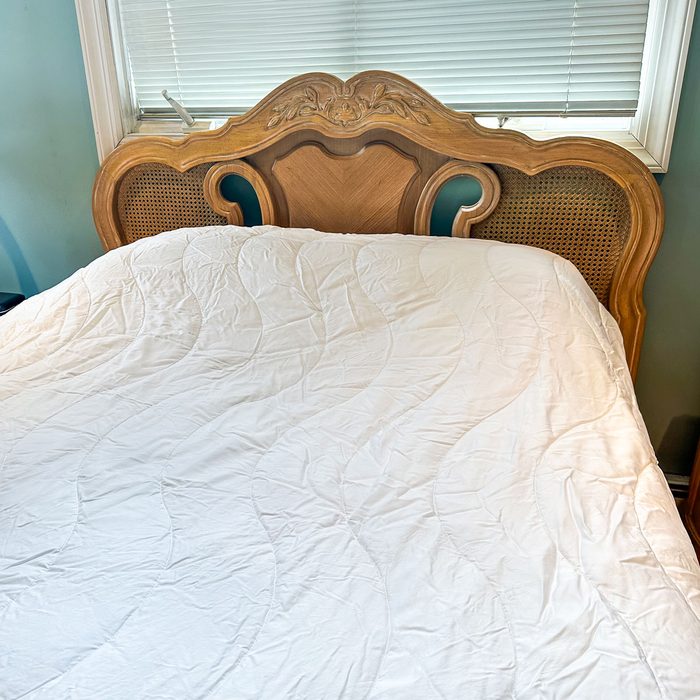Headboard adorns bed with carved details, positioned against closed blinds in a teal-walled room. White quilt covers the mattress smoothly.