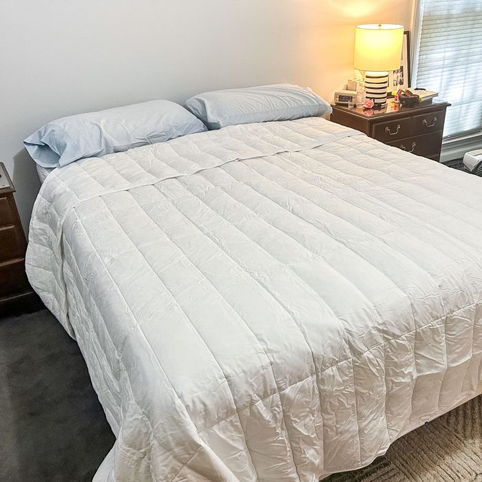 Bed covered with a white quilt; resting under soft lighting from a bedside lamp; two pillows. Bedroom has a nightstand with books and a window.