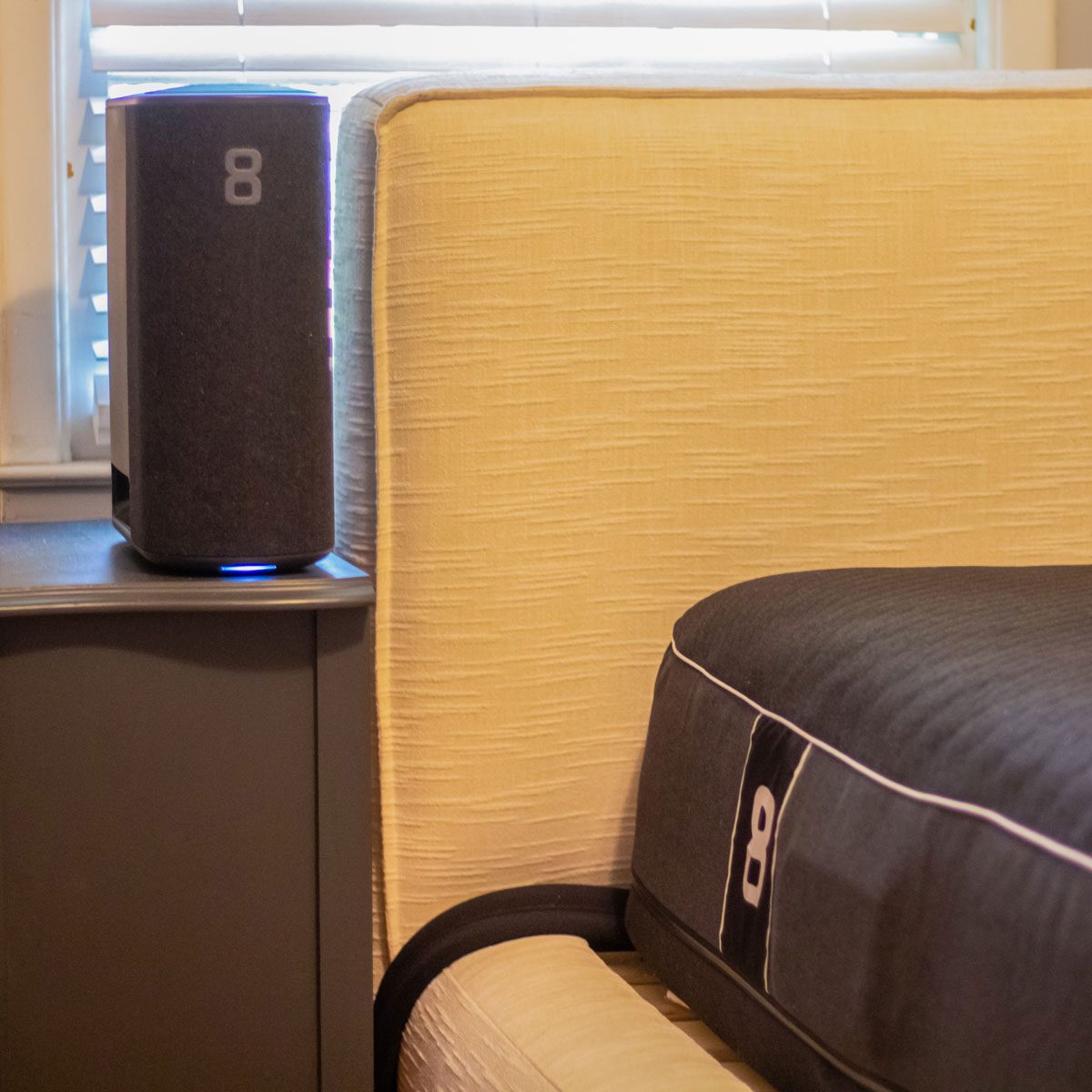 Device displaying "8" rests on a bedside table beside a beige headboard and mattress. Window blinds partially open in the background.