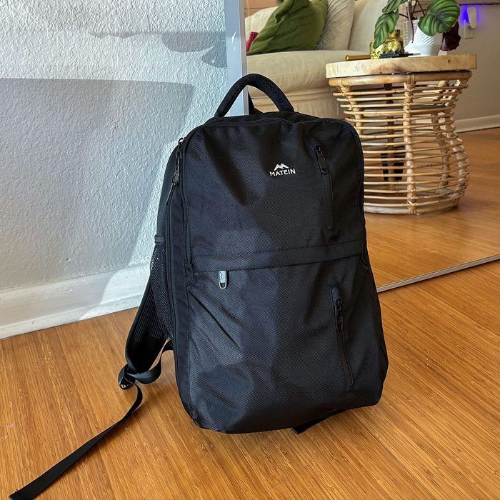 A black backpack stands on a wooden floor, reflecting in a mirror, with a couch and table in the background. Text: 