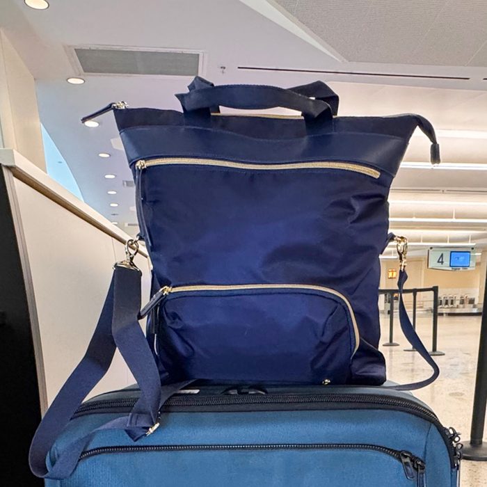 A blue bag rests on a suitcase in an airport setting with check-in counters and screens in the background.