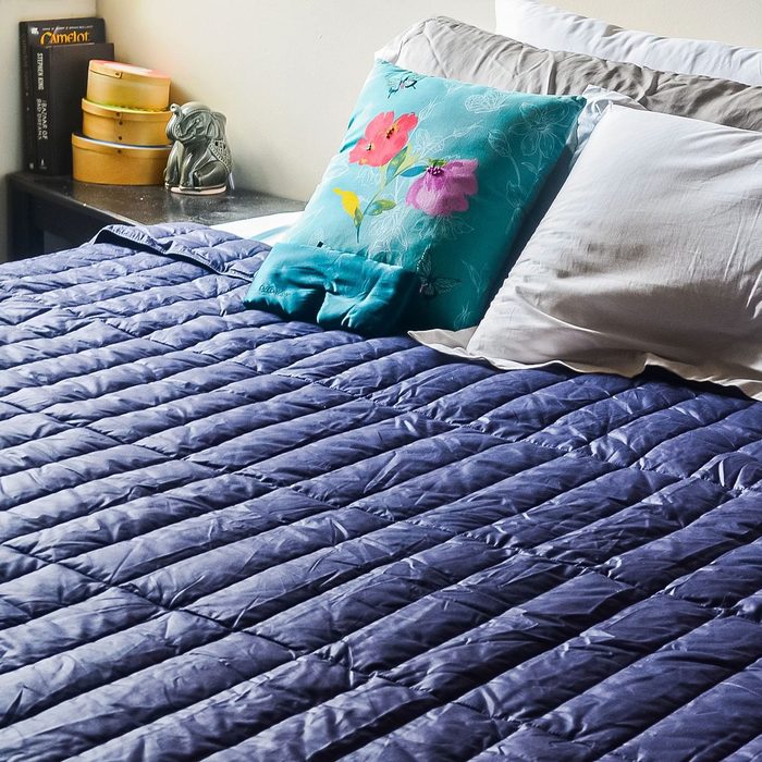 Bed holds floral pillow and white cushions, covered by a blue quilt. A bedside table has books and decorative items.
