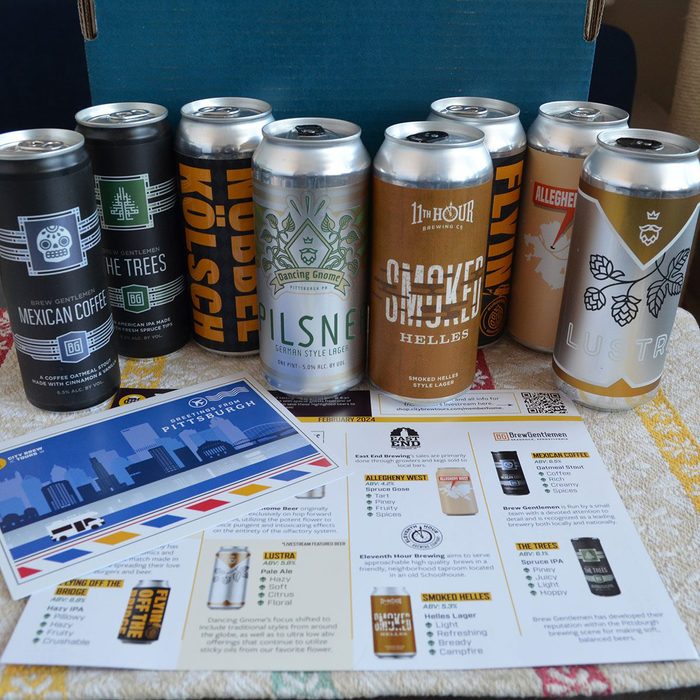 Cans display various beer labels; they stand in two rows. A brochure showcasing beer descriptions and city skyline decorates the table surface.