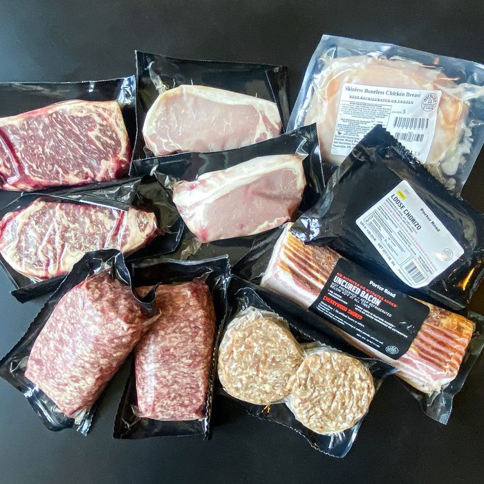 Various packaged meats lie arranged on a dark surface, including beef, pork, bacon, chicken, and sausage patties in clear plastic wrapping.