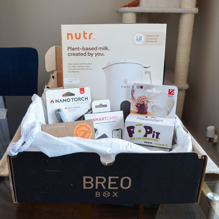 Box holds kitchen gadgets and tools including a NanoTorch, SmartCard, and Popit jar lid remover, placed on a table near a chair.