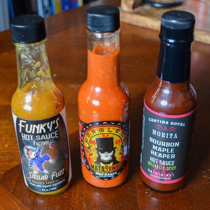 Three hot sauce bottles sit on a wooden table, each featuring colorful labels with unique designs and bold, spicy flavor descriptions.