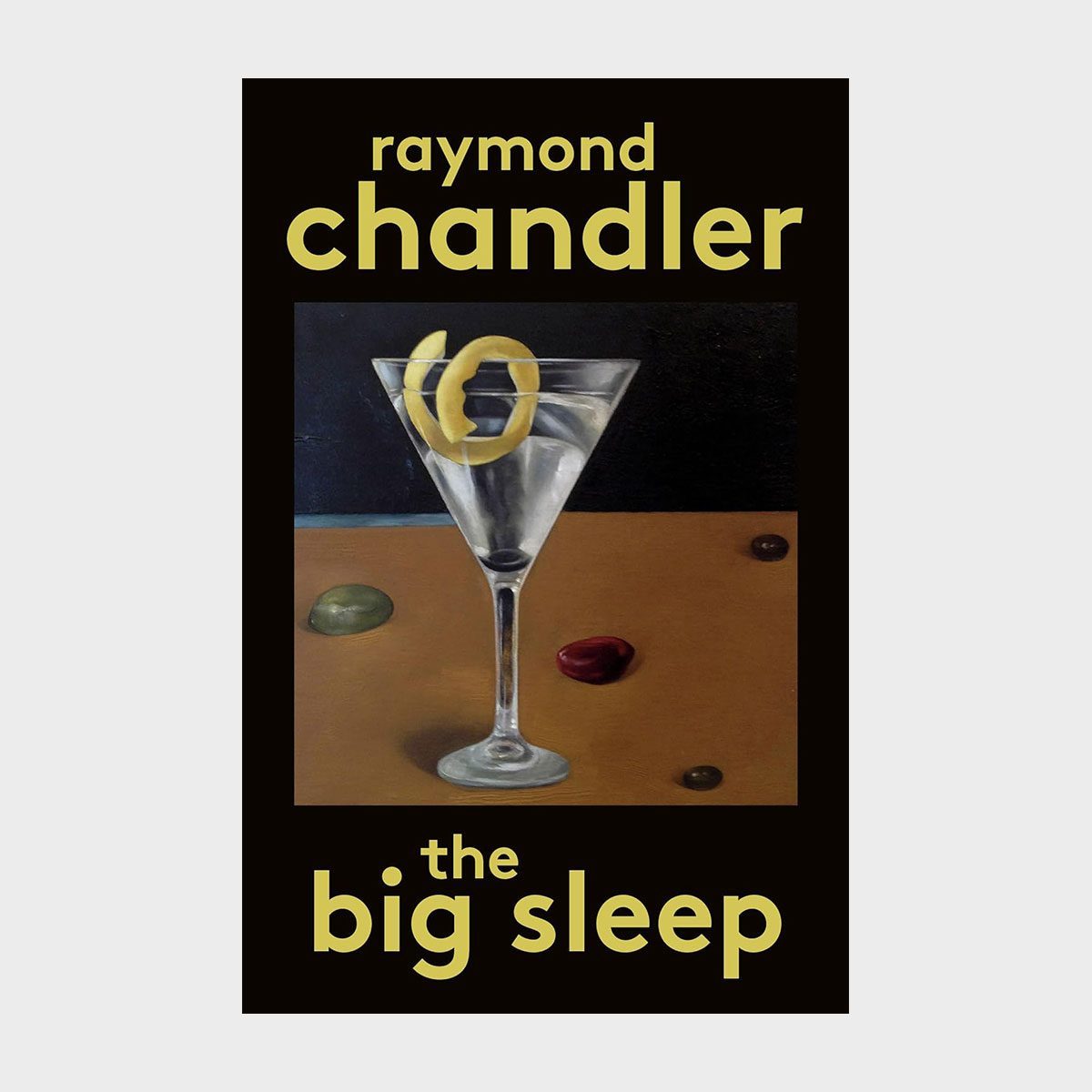 Martini glass holds lemon twist; sits on wooden table with pebbles. Text reads "raymond chandler the big sleep" in yellow on black background.