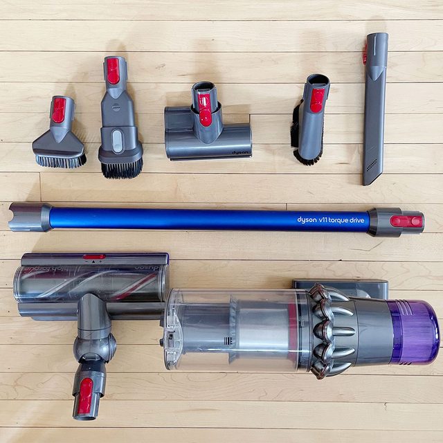 Vacuum cleaner components lying on a wooden floor; includes nozzles, brushes, a tube, and a main unit. Text: 