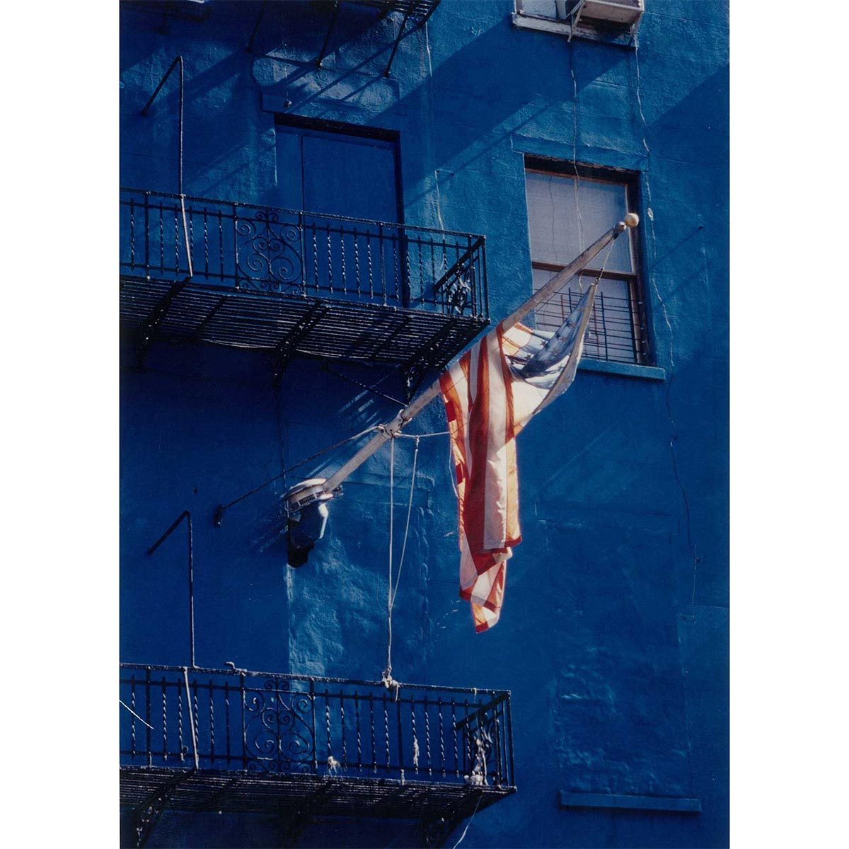 An American flag hangs above blue-painted building, partially draped. Two metal balconies are visible, with shadows cast by sunlight.