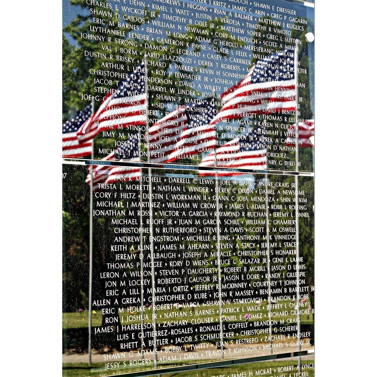 Names etched on a reflective monument; American flags subtly mirrored; trees and sky enhance the patriotic scene.