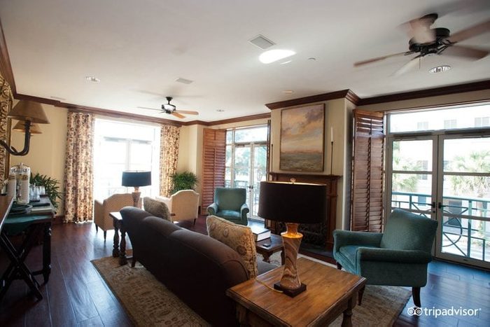 A cozy living room displays furniture arranged around a fireplace, with large windows and floral curtains illuminating the space.