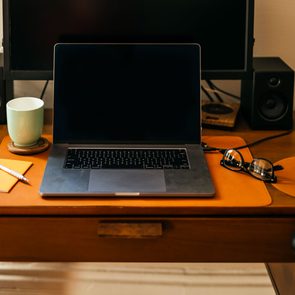 Laptop resting on a wooden desk, surrounded by speakers, a mug, a notebook with a pen, and glasses next to a computer mouse.