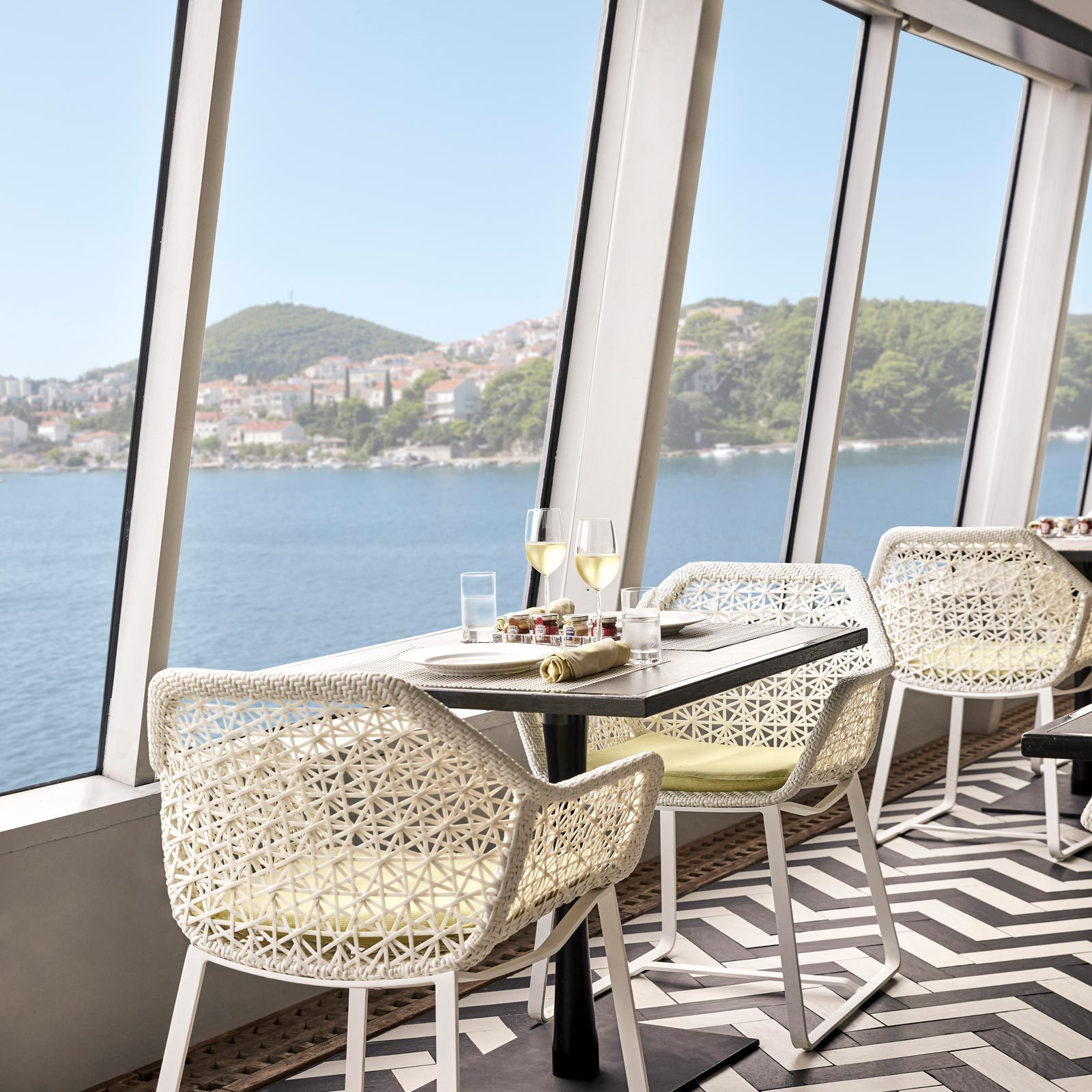 Wicker chairs surround a table with wine glasses, overlooking a window view of a calm sea and distant hills.