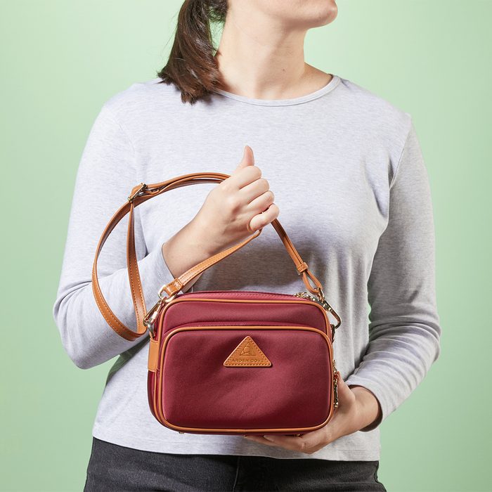 A person holds a small, burgundy handbag with tan straps; they wear a gray top against a light green background.