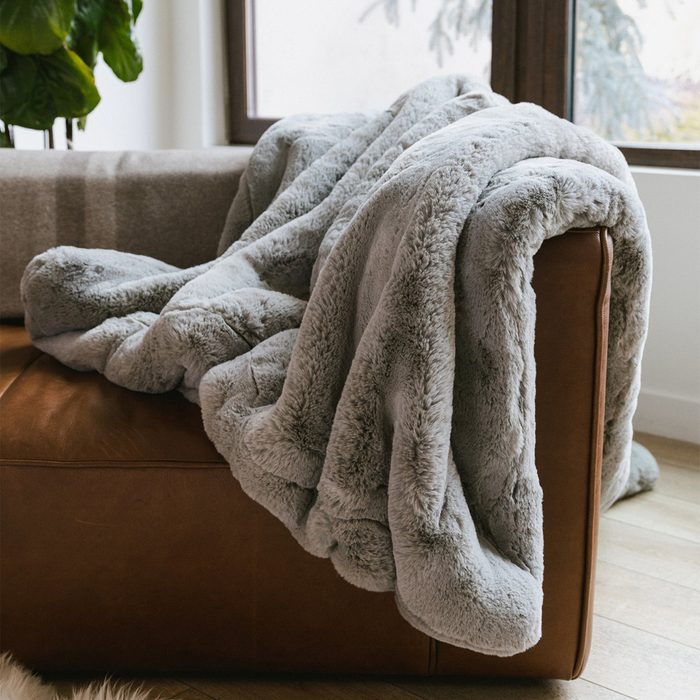 A soft, gray fluffy blanket drapes over a leather sofa, placed next to a window with natural light streaming in.