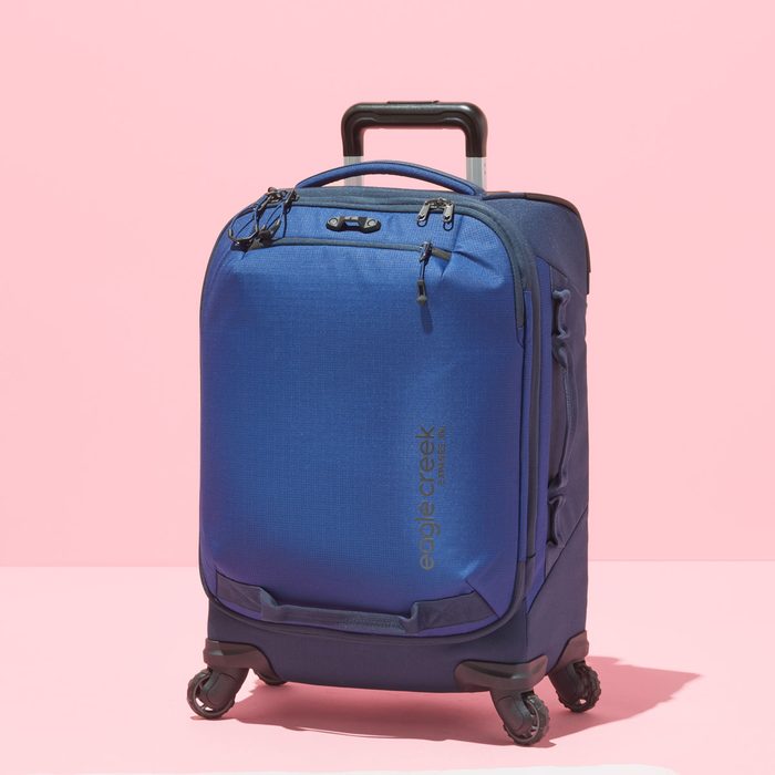 A blue suitcase with wheels and an extended handle sits against a pink backdrop, displaying 