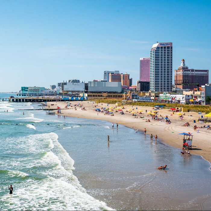 Beachgoers relax and swim alongside gently crashing waves; background features high-rise buildings under a clear blue sky.