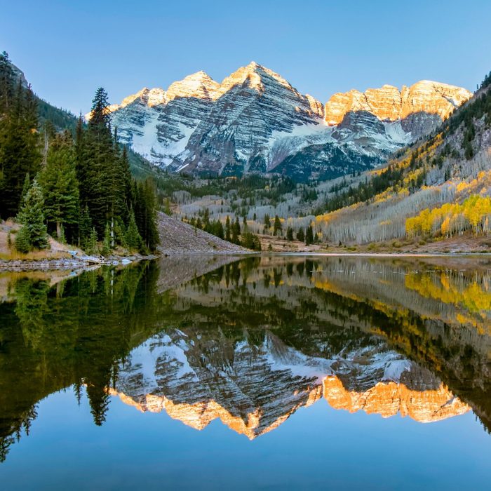 Mountains reflect on a calm lake, surrounded by pine trees and autumn foliage under a clear blue sky.