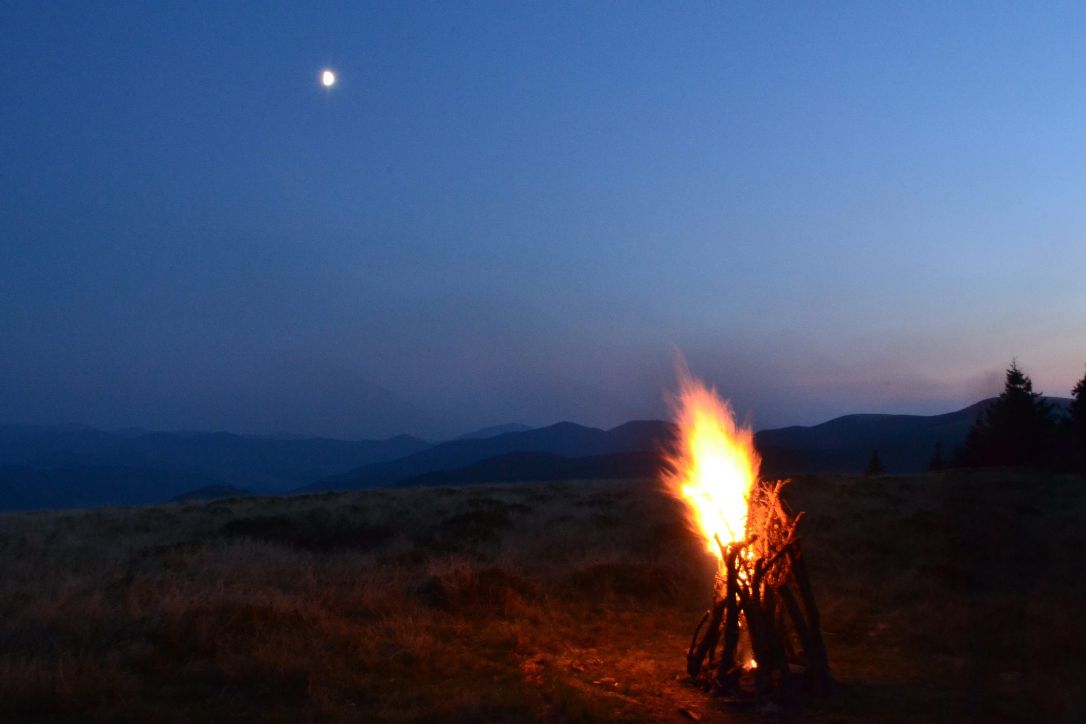 A campfire burns brightly on a grassy hillside at dusk, with mountains silhouetted in the background under a moonlit sky.