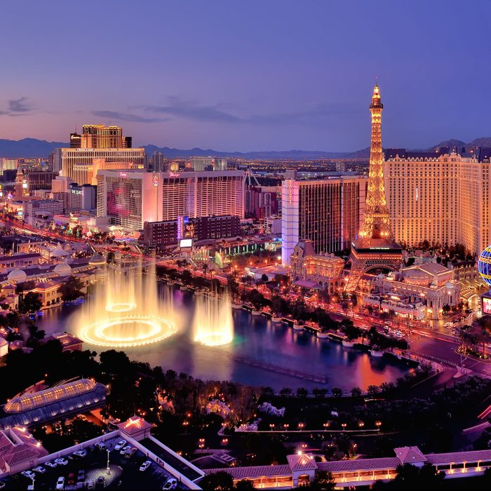 Fountains perform water show amid illuminated cityscape featuring tall buildings and replica Eiffel Tower under evening sky.