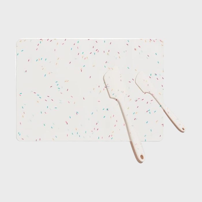 Cutting board lies flat, patterned with colorful sprinkles, surrounded by two spatulas matching the design, on a plain light background.