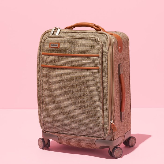 Suitcase stands upright, featuring textured fabric with brown leather trim, wheels, and handles; set against a solid pink background.