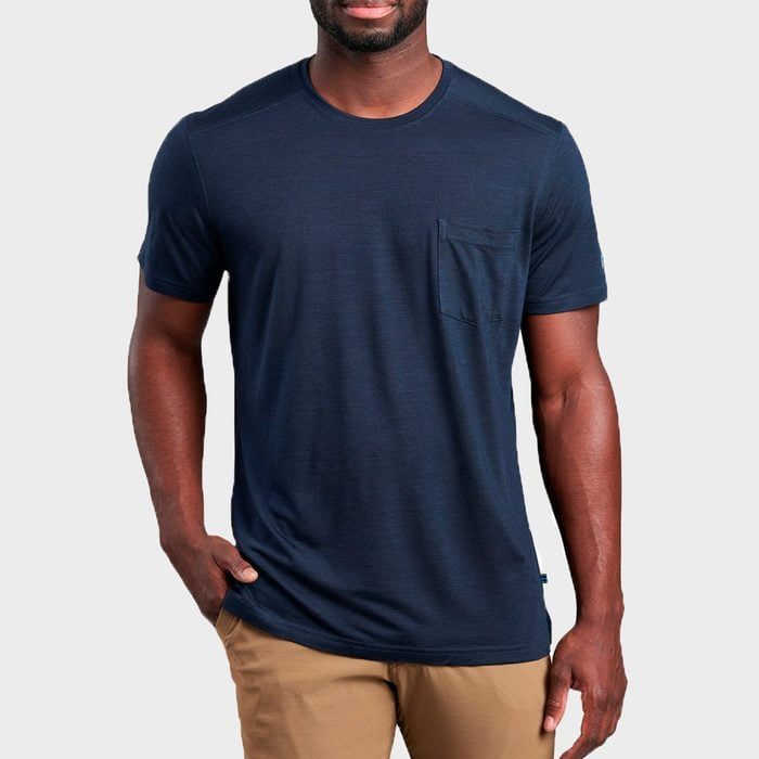 A person stands wearing a navy T-shirt with a small chest pocket and tan pants against a plain background.