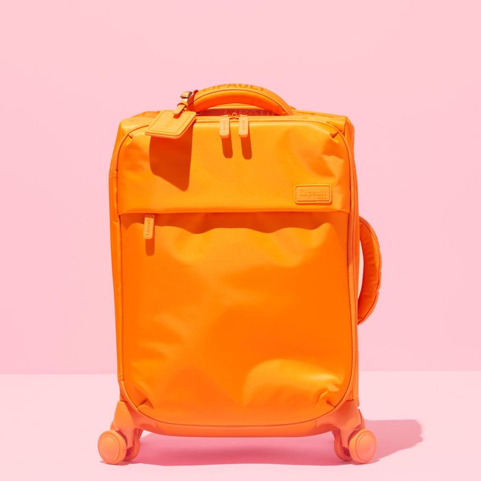 Orange suitcase stands still on four wheels against a pink background, featuring a fabric tag and visible zippers.