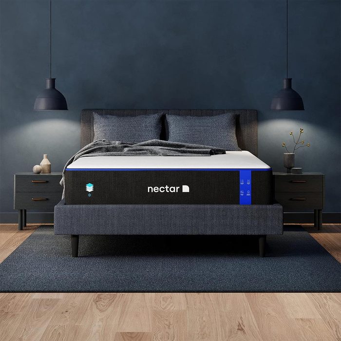 A bed rests in a dimly lit room, flanked by minimalist nightstands, beneath matching wall-mounted lamps. The mattress brand, 
