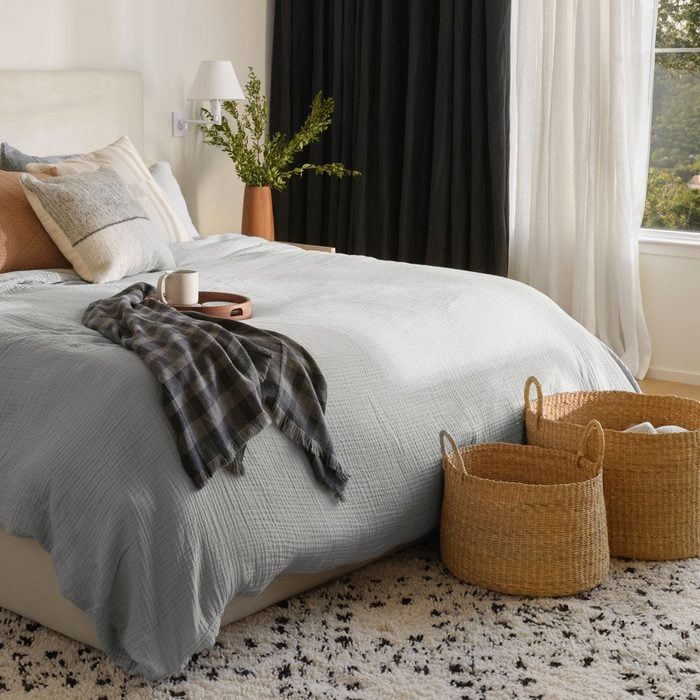 Bed with plaid cloth and mug, in a cozy bedroom with green vase, wicker baskets, and soft lighting from curtains.