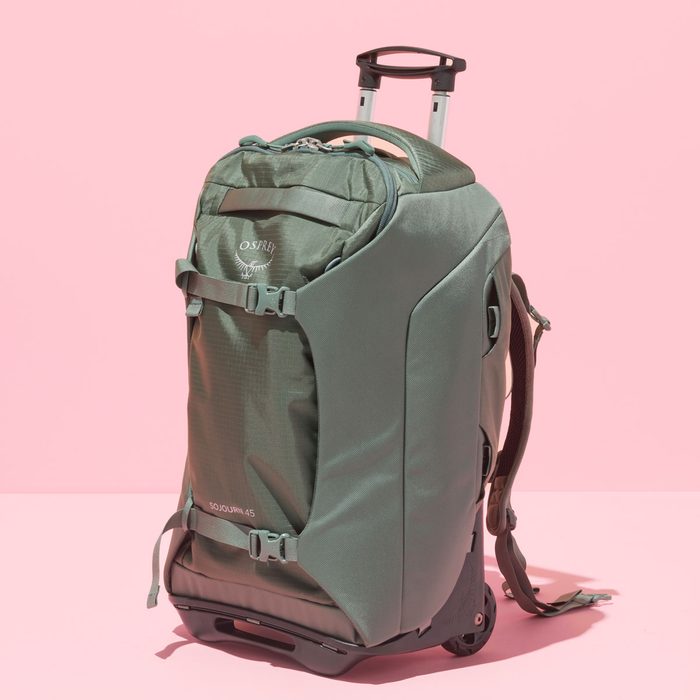 Green wheeled backpack stands upright against a pink background. 