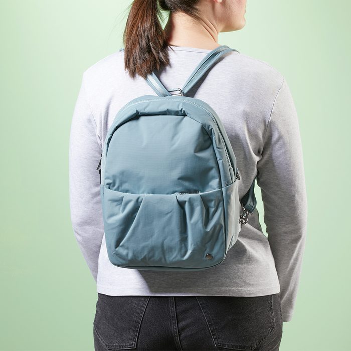 A person wearing a light blue backpack stands against a pale green background, dressed in a gray shirt and dark pants.