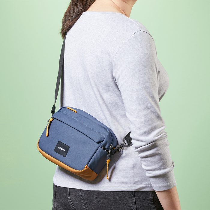 A person wears a blue crossbody bag with tan accents over a gray shirt, standing against a green background.